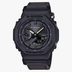 A black CASIO G-SHOCK watch with a digital display and analog hands, featuring a carbon case and a black cloth band.